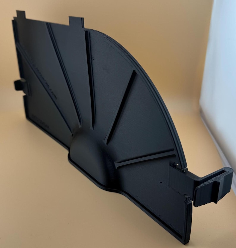 May include: A black, fan-shaped plastic component with a curved edge and several angled ridges. The piece has a semi-circular base and a rectangular section with tabs. The object is likely a part of a larger assembly.