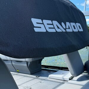 Puede incluir: Se ve una lona de barco gris oscuro con el texto blanco "SEA DOO". La lona est&aacute; colocada sobre un asiento de barco. El interior del barco es gris y el fondo muestra un lago y &aacute;rboles.