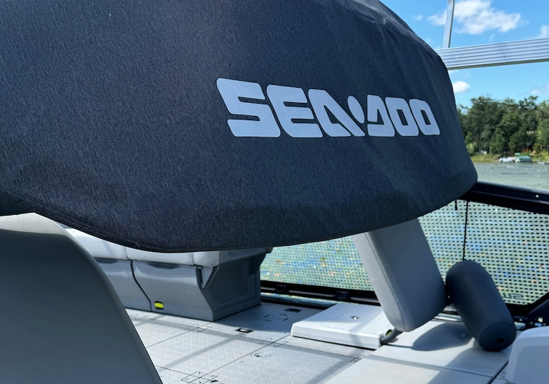 Puede incluir: Se ve una lona de barco gris oscuro con el texto blanco "SEA DOO". La lona est&aacute; colocada sobre un asiento de barco. El interior del barco es gris y el fondo muestra un lago y &aacute;rboles.