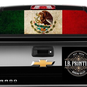 May include: A black pickup truck with a Mexican flag decal on the rear window. The truck has a "SILVERADO" emblem on the tailgate and a sticker with the text "I.B. PRINTING EST. 2024 MORENO VALLEY" on the right side of the tailgate.