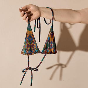 Può includere: Un colorato top bikini a triangolo con un motivo geometrico nei toni del blu, arancione, giallo e verde. Il top ha spalline annodabili e un fiocco al centro. Il top del bikini è tenuto da una mano.