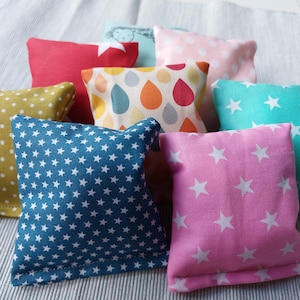 Könnte beinhalten: Eine Sammlung kleiner, quadratischer Stoffkissen in verschiedenen Farben und Mustern. Designs umfassen weiße Sterne auf Blau und Rosa, Polka Dots und bunte Tropfen. Die Kissen sind auf einer hellgrau gestreiften Oberfläche angeordnet.
