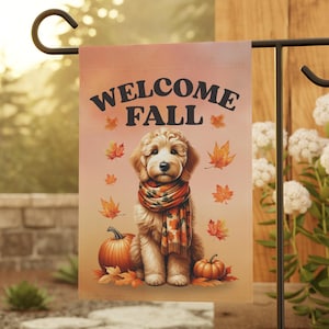 May include: A garden flag with the text "WELCOME FALL" features a golden doodle wearing a scarf, surrounded by pumpkins and autumn leaves. The flag is displayed on a black metal stand, perfect for fall decor.