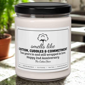 May include: A white candle in a glass jar with a black lid. The label reads "smells like COTTON, CUDDLES & COMMITMENT" and "Happy 2nd Anniversary". The candle is on a countertop with a plant in the background.