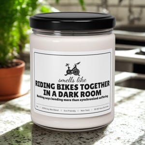 May include: A white candle in a glass jar with a black lid. The label reads "smells like Riding Bikes Together in a Dark Room." The candle has a cycling graphic and is eco-friendly.