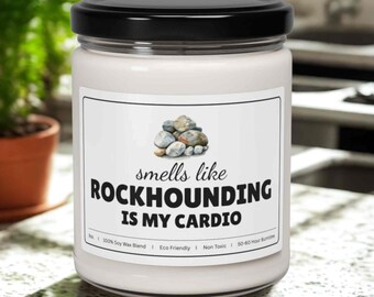 May include: A white candle in a glass jar with a black lid. The label reads "smells like ROCKHOUNDING IS MY CARDIO" with a graphic of rocks. The candle is on a speckled countertop with a plant in the background.
