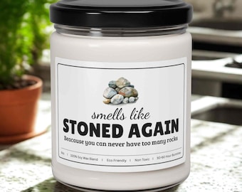 May include: A white candle in a glass jar with a black lid. The label reads "smells like STONED AGAIN" with a graphic of stones. The text below says "Because you can never have too many rocks."