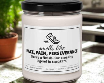 May include: A white candle in a glass jar with a black lid. The label features a running shoe graphic and the text "smells like PACE, PAIN, PERSEVERANCE. You're a finish-line-crossing legend in sneakers."