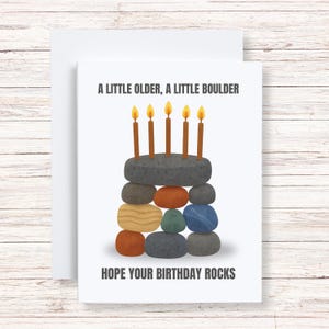 May include: A white birthday card with the text "A LITTLE OLDER, A LITTLE BOULDER HOPE YOUR BIRTHDAY ROCKS." The card features a stacked stone cake with five lit candles. The cake is made of various colored stones.