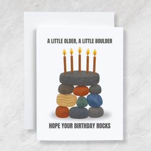 May include: A white birthday card with the text "A LITTLE OLDER, A LITTLE BOULDER" and "HOPE YOUR BIRTHDAY ROCKS." The card features a stack of colorful stones with five lit candles on top, resembling a cake.