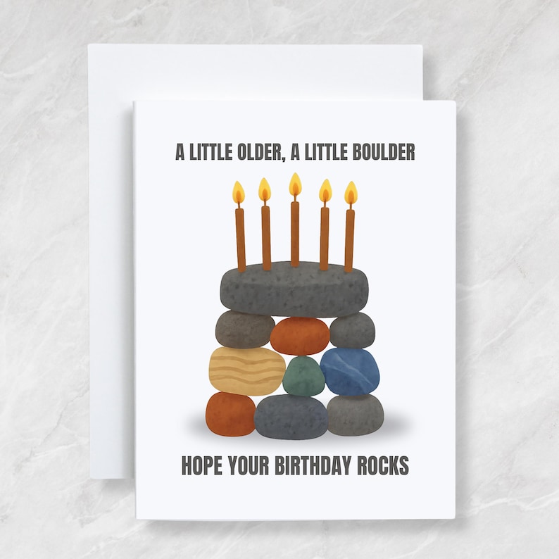 May include: A white birthday card with the text "A LITTLE OLDER, A LITTLE BOULDER" and "HOPE YOUR BIRTHDAY ROCKS." The card features a stack of colorful stones with five lit candles on top, resembling a cake.