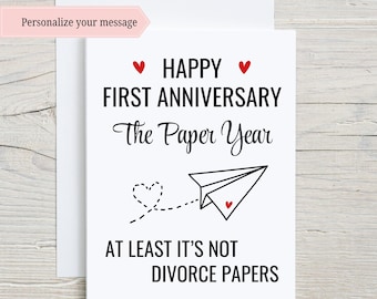 May include: A white card with a red heart and the text "Happy First Anniversary The Paper Year" and a paper airplane with a red heart. The text below the airplane says "At least it's not divorce papers."