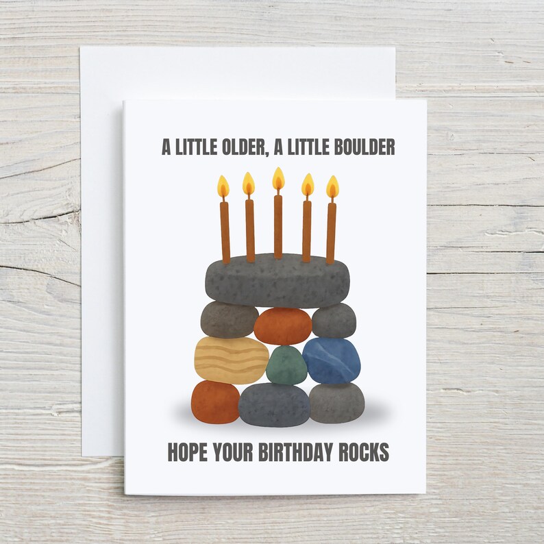 May include: A birthday card with a unique design. The card features a stack of colorful rocks arranged to resemble a cake, topped with five lit candles. Text on the card reads: "A LITTLE OLDER, A LITTLE BOULDER. HOPE YOUR BIRTHDAY ROCKS."