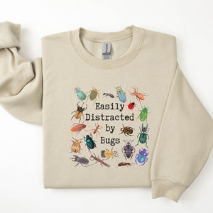 May include: A beige sweatshirt with a circle of colourful bug illustrations and the text "Easily Distracted by Bugs".