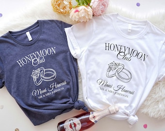 May include: Two navy blue and white t-shirts with the text "Honeymoon Club" and "Maui Hawaii" with a graphic of two wedding rings. The text "MR & MRS JOHNSON" is printed below the graphic.