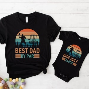 Puede incluir: Dos camisetas negras con un gráfico retro de una persona jugando al disco golf. La camiseta más grande dice "Best Dad By Par" y la más pequeña dice "Disc Golf Caddy".