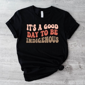 May include: Black t-shirt with the text "It's a good day to be Indigenous" in a retro font with pink, orange, and brown colors.