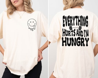 May include: A white oversized t-shirt with a black dripping smiley face on the front and the text "EVERYTHING HURTS AND I'M HUNGRY" on the back.