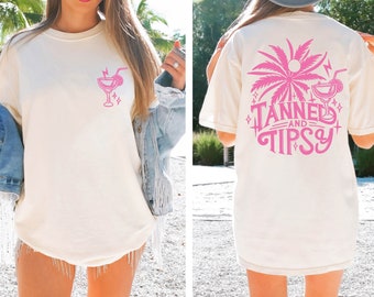 May include: A white t-shirt with a pink graphic of a palm tree and a cocktail glass on the back. The front of the shirt has a pink graphic of a cocktail glass. The text on the back of the shirt reads "Tanned and Tipsy".