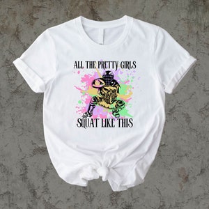 May include: White t-shirt with a colorful graphic of a softball player in a catcher's stance. The text "All the pretty girls squat like this" is printed on the shirt.