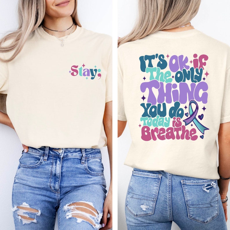 Pode incluir: Uma camiseta bege claro com o texto "Stay;" na frente e "It's ok if the only thing you do today is breathe" nas costas. O texto est&aacute; em uma fonte retr&ocirc; colorida com detalhes roxos, azuis e verdes.