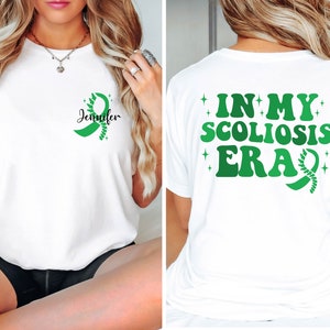 Pu&ograve; includere: Maglietta bianca con un nastro verde e il testo "Jennifer" sul davanti. La parte posteriore della maglietta ha il testo "In My Scoliosis Era" con un nastro verde.