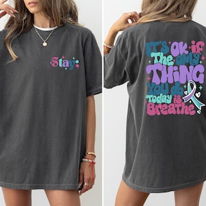 Pode incluir: Uma camiseta cinza escura de tamanho grande com um gr&aacute;fico colorido nas costas que diz "It's ok if the only thing you do today is breathe" e um gr&aacute;fico menor na frente que diz "Stay."