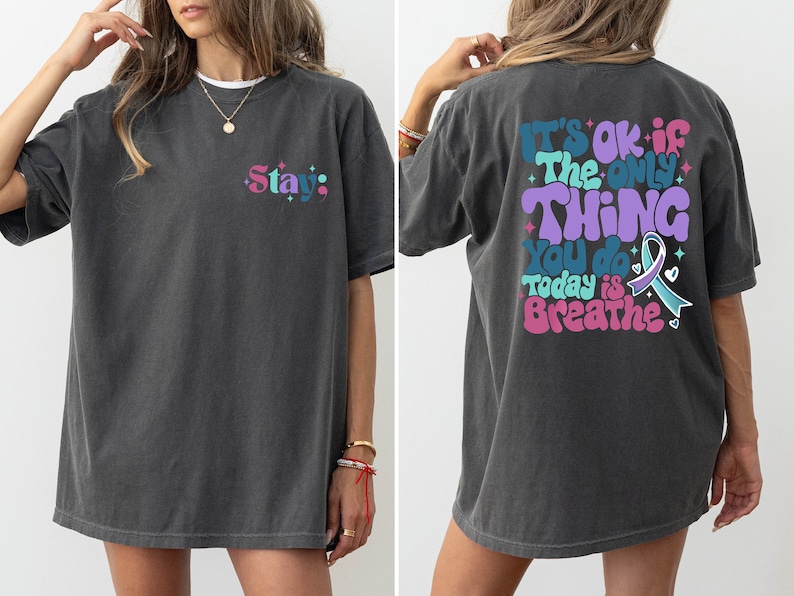 Pode incluir: Uma camiseta cinza escura de tamanho grande com um gr&aacute;fico colorido nas costas que diz "It's ok if the only thing you do today is breathe" e um gr&aacute;fico menor na frente que diz "Stay."