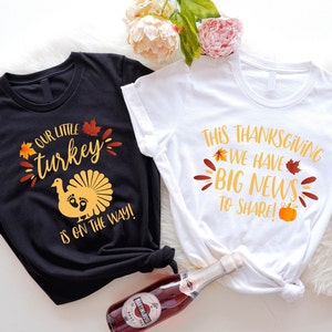 May include: Two t-shirts, one black and one white, with Thanksgiving-themed text. The black shirt says "Our little turkey is on the way!" with a turkey graphic. The white shirt says "This Thanksgiving we have big news to share!" with fall leaves.