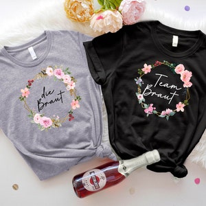 May include: Two t-shirts, one gray and one black, with floral wreath designs. The gray shirt says "die Braut" and the black shirt says "Team Braut". Both shirts have a round neck and short sleeves. A bottle of sparkling wine is in the foreground.