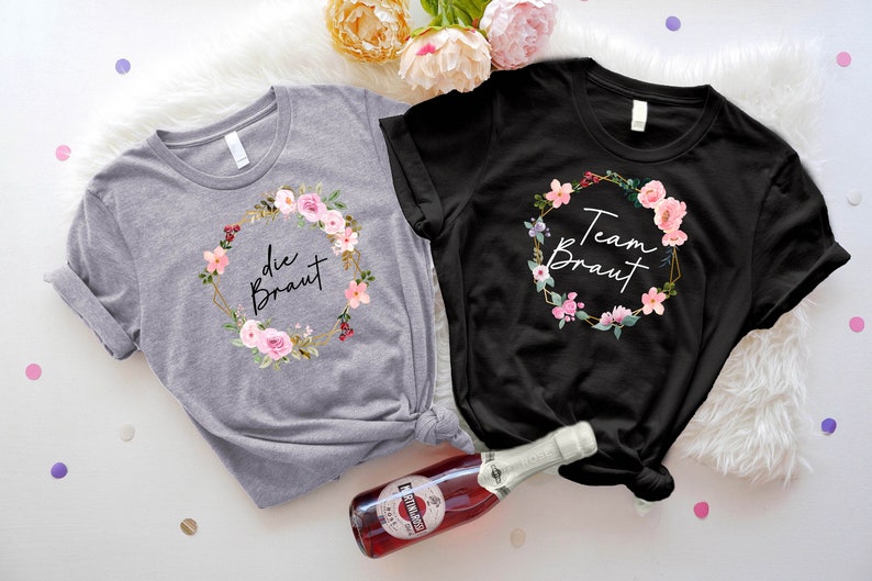 May include: Two t-shirts, one gray and one black, with floral wreath designs. The gray shirt says "die Braut" and the black shirt says "Team Braut". Both shirts have a round neck and short sleeves. A bottle of sparkling wine is in the foreground.