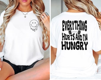 May include: White t-shirt with a black dripping smiley face on the front and the text "EVERYTHING HURTS AND I'M HUNGRY" with a barbell graphic on the back.