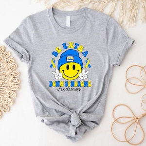 Op de afbeelding: Grijs T-shirt met een blauwe en gele smiley die een blauwe pet draagt met de letters "T21" erop. De tekst "Be Extra Down Syndrome Awareness" staat onder de smiley.