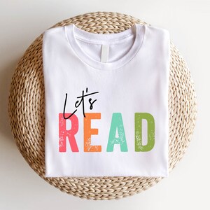 Puede incluir: Camiseta blanca con el texto "Let's Read" en letras coloridas y desgastadas. Las letras son en tonos de rosa, naranja, verde y azul.