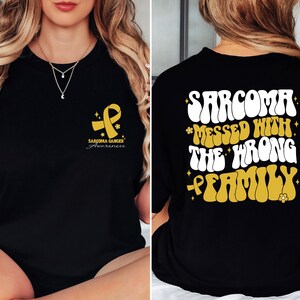 Op de afbeelding: Zwart T-shirt met een gouden lint en de tekst "Sarcoma Cancer Awareness" op de voorkant. De achterkant van het shirt heeft de tekst "Sarcoma *Messed With* The *Wrong* Family" in een retro-lettertype met gouden accenten.