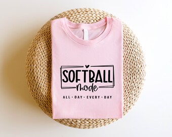 Op de afbeelding: Een roze T-shirt met de tekst "Softball Mode" in zwarte letters. De tekst is omlijst in een zwart rechthoek met een klein hartje erboven. Onder de rechthoek staat de tekst "All Day Every Day".