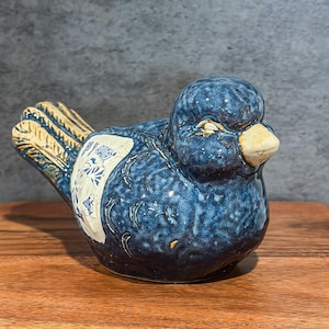 May include: A ceramic bird figurine in shades of blue, with a cream-colored beak and accents. The bird has a textured surface and a decorative patch with a floral design. The figurine is displayed on a wooden surface.