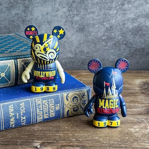 May include: Two Disney Bearbrick figures. One is navy blue with yellow accents and reads "Hollywood Studios 1989." The other is navy blue with red accents and reads "Magic Kingdom 1971."