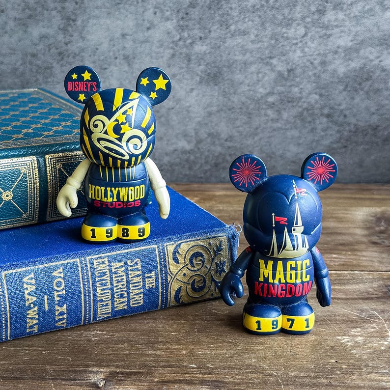 May include: Two Disney Bearbrick figures. One is navy blue with yellow accents and reads "Hollywood Studios 1989." The other is navy blue with red accents and reads "Magic Kingdom 1971."
