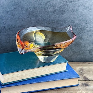 May include: A clear glass bowl with an organic, asymmetrical shape featuring a gradient of amber, yellow, and orange hues. The bowl has a smooth, polished surface and a thick base, showcasing a translucent quality that catches the light.