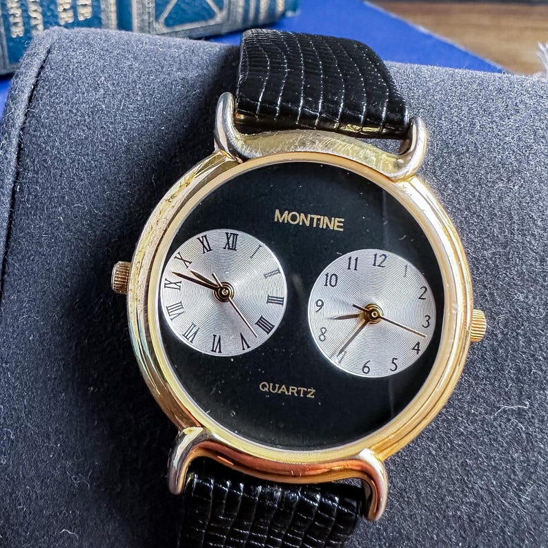 May include: A Montine quartz wristwatch with a gold-tone round case and a black textured leather strap. The black dial features two silver-toned sub-dials, one with Roman numerals and one with Arabic numerals, each equipped with gold-toned hands.