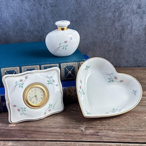 May include: A white porcelain set featuring a round perfume bottle, a rectangular desk clock with a gold-tone bezel and Roman numerals, and a heart-shaped dish. Each piece is decorated with a delicate pattern of pink rosebuds and green leaves.