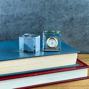May include: Two clear glass cubes resting on a blue book. One cube features a circular analog clock face with a gold-tone bezel, black hands, and a small red and white flag icon at the twelve o'clock position. The other cube is plain.
