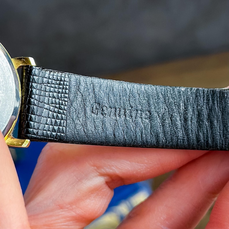 May include: A black watch strap with a textured surface and the word Genuine embossed on the inner side. The strap is attached to a gold-toned metal watch case. The material features a fine grain pattern near the attachment point.
