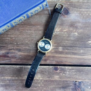 May include: A Montine quartz wristwatch featuring a round gold-tone case with a black dial. The dial displays two smaller silver-tone sub-dials with black numerals. The watch is fitted with a black textured leather strap and a gold-tone buckle.