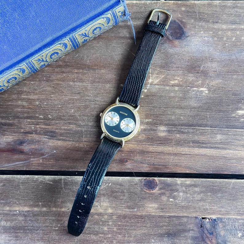 May include: A Montine quartz wristwatch featuring a round gold-tone case with a black dial. The dial displays two smaller silver-tone sub-dials with black numerals. The watch is fitted with a black textured leather strap and a gold-tone buckle.