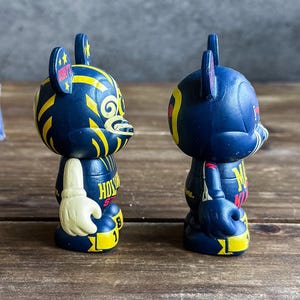 May include: Two navy blue bear-shaped figurines with yellow and white detailing. One has a white glove and the word "HOLLYWOOD" on its chest. The other has the letter "M" on its chest.