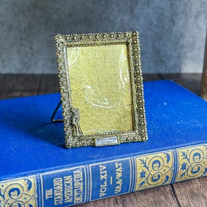 May include: A vintage gold-toned metal picture frame with a decorative beaded border. The frame has a small stand and features a couple embracing at the bottom. The text "UNIVERSAL STUDIOS CALIFORNIA" is visible. The frame is resting on a blue book.