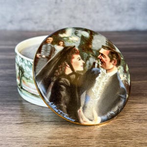 May include: A round ceramic trinket box featuring a lid with a painted scene of a couple in formal attire, with the woman wearing a black veil. The side of the box shows a landscape illustration. The signature Lillian T. Chambers is visible.