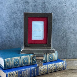 May include: A silver-toned metal stocking holder featuring a rectangular frame with a deep red fabric border. The center insert displays the text Pottery Barn, grosgrain frame stocking holder, and 2 x 3 inches / 5 x 8 cm. A metal hook extends from the base.